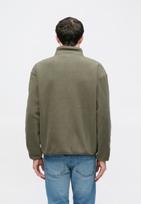 Άνδρας με πλάτη φορώντας πράσινο χακί φλις μπουφάν και μπλε τζιν μπροστά σε λευκό φόντο.