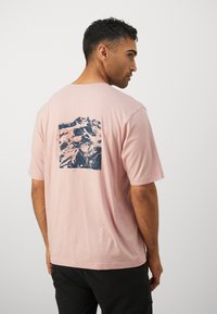 T-shirt a maniche corte rosa chiaro con una grafica di montagne blu e rosa sul retro. Tessuto morbido, vestibilità comoda e scollatura classica a girocollo.