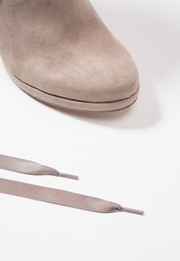 Close-up of a beige suede shoe toe next to two loose taupe satin shoelaces on a white background.