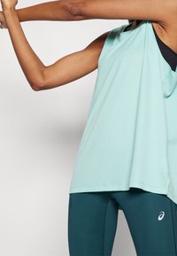 Top sin mangas verde menta con un corte relajado y aberturas laterales, combinado con leggings azul oscuro que presentan un logo en la parte inferior de la pierna.