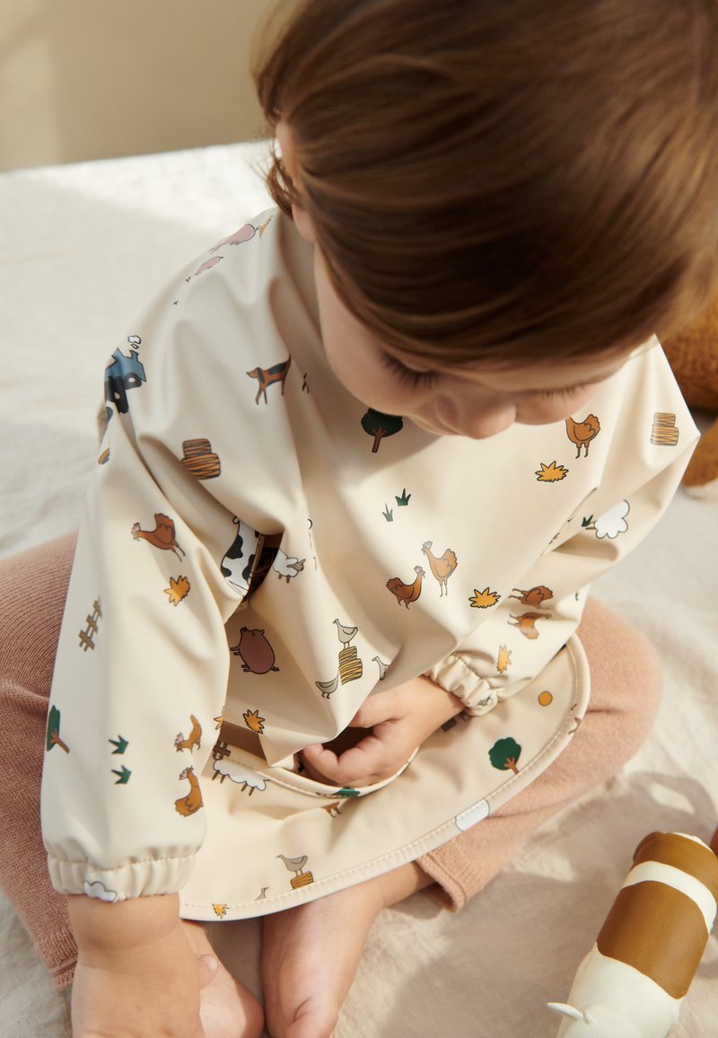 Beige long-sleeved children's smock with colorful farm animal print. Features elastic cuffs and a rounded hem, textured soft fabric.