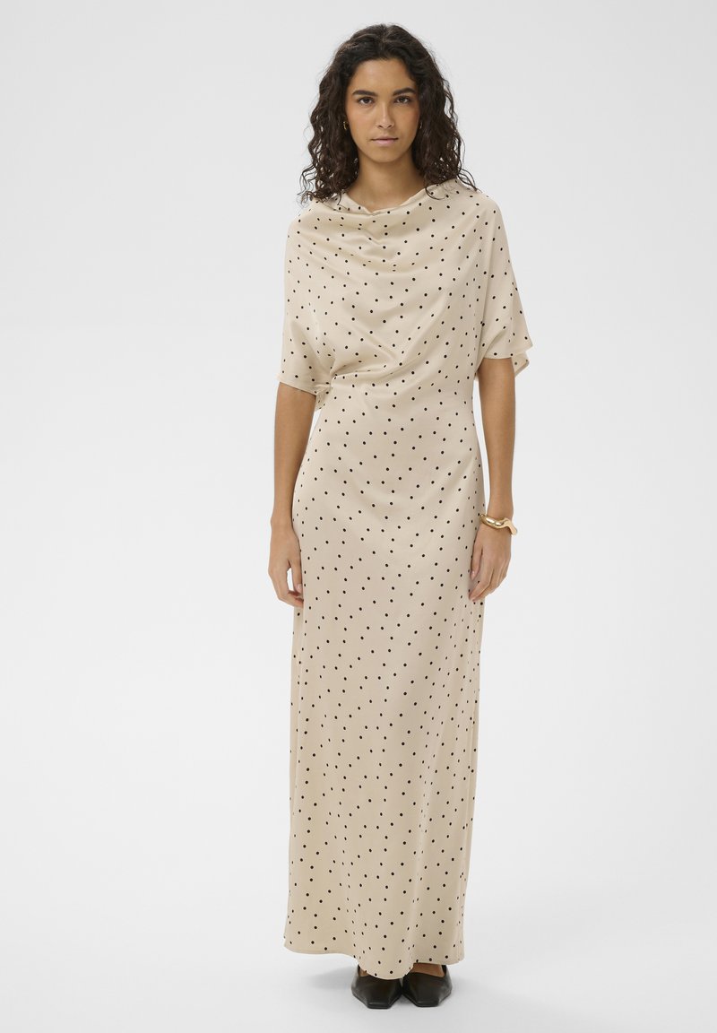 Vrouw draagt een beige, enkellange jurk met zwarte polkadots, korte mouwen, een gedrapeerde halslijn, zwarte schoenen en een gouden armband.