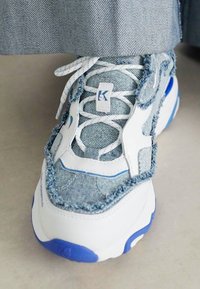 Bílé a modré tenisky z denimu s roztrhanými okraji, nošené s modrými džínami.