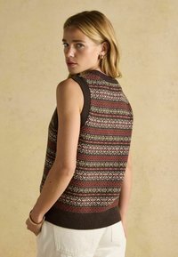 Ženska z blond lasmi nosi brezrokavnik iz pletene tkanine v rjavi, rdeči in beli barvi, v kombinaciji s svetlimi hlačami, obrnjena proč z stranskim pogledom.