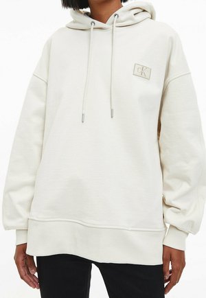 Mujer que lleva una sudadera holgada con capucha de color blanco roto con cordones y un pequeño logo de CK en el pecho y el puño, combinada con pantalones negros.