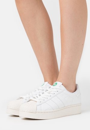 Zapatillas deportivas blancas con plataforma, punteras texturizadas y logo verde en la lengüeta, usadas por una persona de pie sobre un fondo blanco.