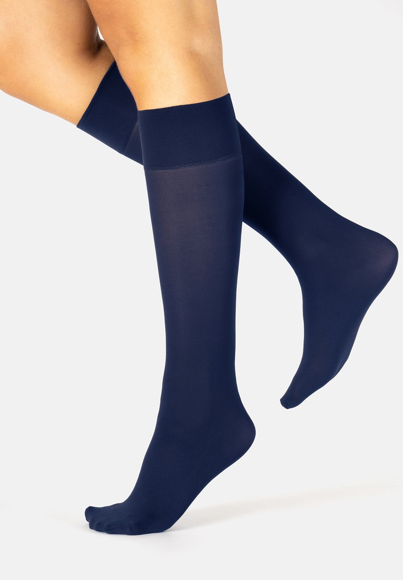 Piernas con calcetines de compresión oscuros hasta la rodilla, un pie plano y el otro ligeramente levantado sobre los dedos contra un fondo blanco.