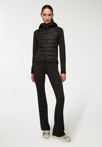 Czarna pikowana kurtka z kapturem, z zapinaniem na zamek z przodu i dopasowanym krojem, w zestawie z jednolitymi czarnymi legginsami i wielokolorowymi sneakersami.