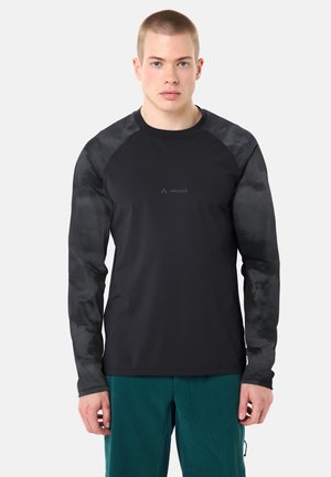 Junger Mann mit kurzen Haaren, der ein schwarzes Langarmshirt mit Camouflage-Ärmeln und grüne Hose trägt, vor einem schlichten hellen Hintergrund stehend.