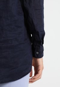 Marineblauw linnen overhemd met een gestructureerde afwerking, lange mouwen, met knopen afgewerkte manchetten en een relaxed fit, weergegeven van achteren met een hand die het vasthoudt.