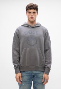 Hanorac gri cu buzunar frontal, prezentând un logo „BOSS” ridicat și un grafic cu munte. Material moale, texturat, croială lejeră și glugă cu șnur.
