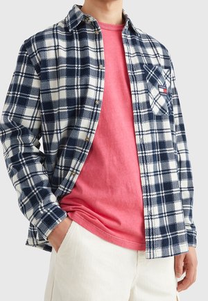 Mand iført marineblå og hvid ternet skjorte over en pink t-shirt og hvide bukser med én hånd i lommen mod en ensfarvet baggrund.