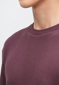 Bordowy sweter z dzianiny ze wzorem fakturowanym, z okrągłym dekoltem i widocznym przeszyciem przy kołnierzu. Ukazana szyja modela bez określonych cech.