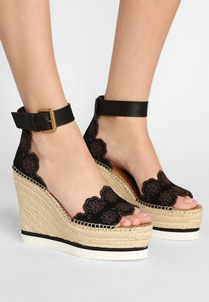 Des sandales à talon noir présentent un design floral découpé, des sangles en cuir avec une fermeture à boucle, et une plateforme en jute. Texturées et ouvertes au niveau des orteils.