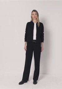 Zwarte bomberjack met ritssluiting over een witte top, gecombineerd met zwarte broek. De outfit kenmerkt zich door gladde texturen en een losse pasvorm. Zwarte schoenen maken de look af.