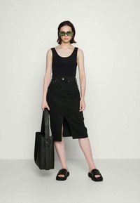 Top preto de alças, saia de ganga preta de cintura alta com fenda frontal, chinelos pretos e uma bolsa de tote preta. Óculos de sol verdes acrescentam um toque de cor.