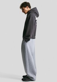 Temno siva hoodie s kapuco in sprednjim žepom, v kombinaciji s svetlo sivo ohlapnimi trenirkami. Obe sta izdelani iz mehkega materiala. Črne superge.