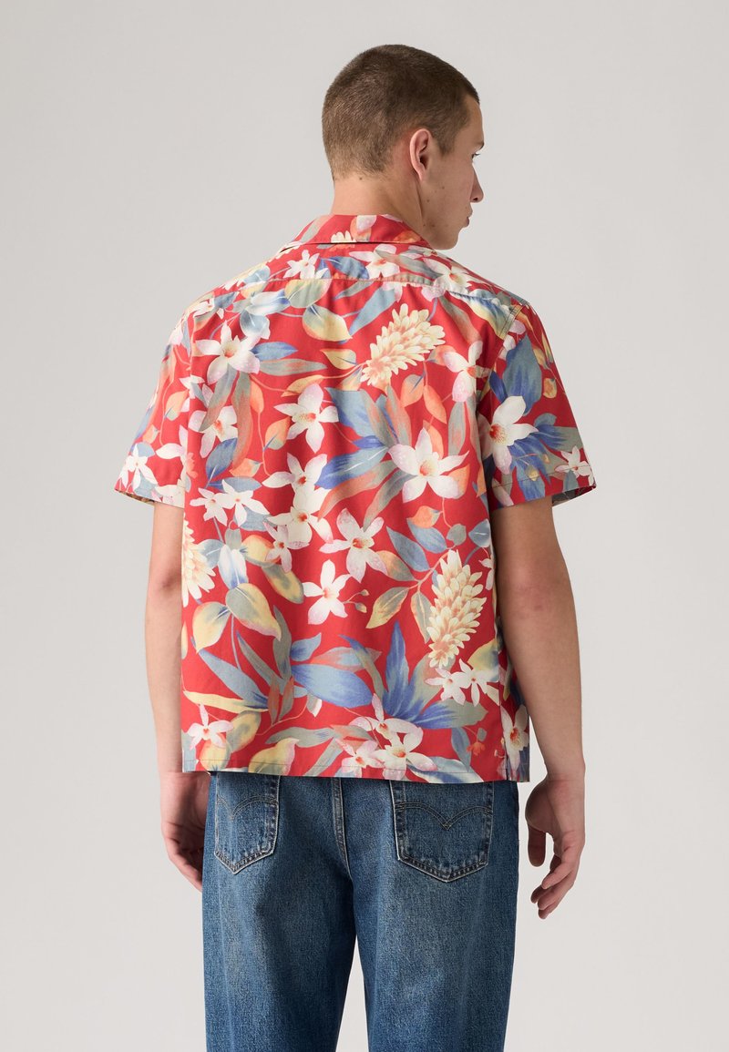 Hombre de espaldas vistiendo una camisa roja de manga corta con motivos florales blancos, azules y amarillos y vaqueros azules.