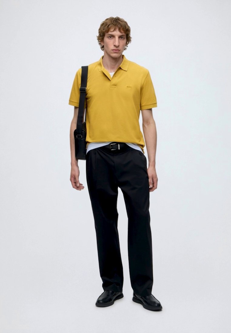 Jeune homme debout portant un polo jaune moutarde, pantalon noir, ceinture noire, chaussures noires et un sac d'épaule noir.