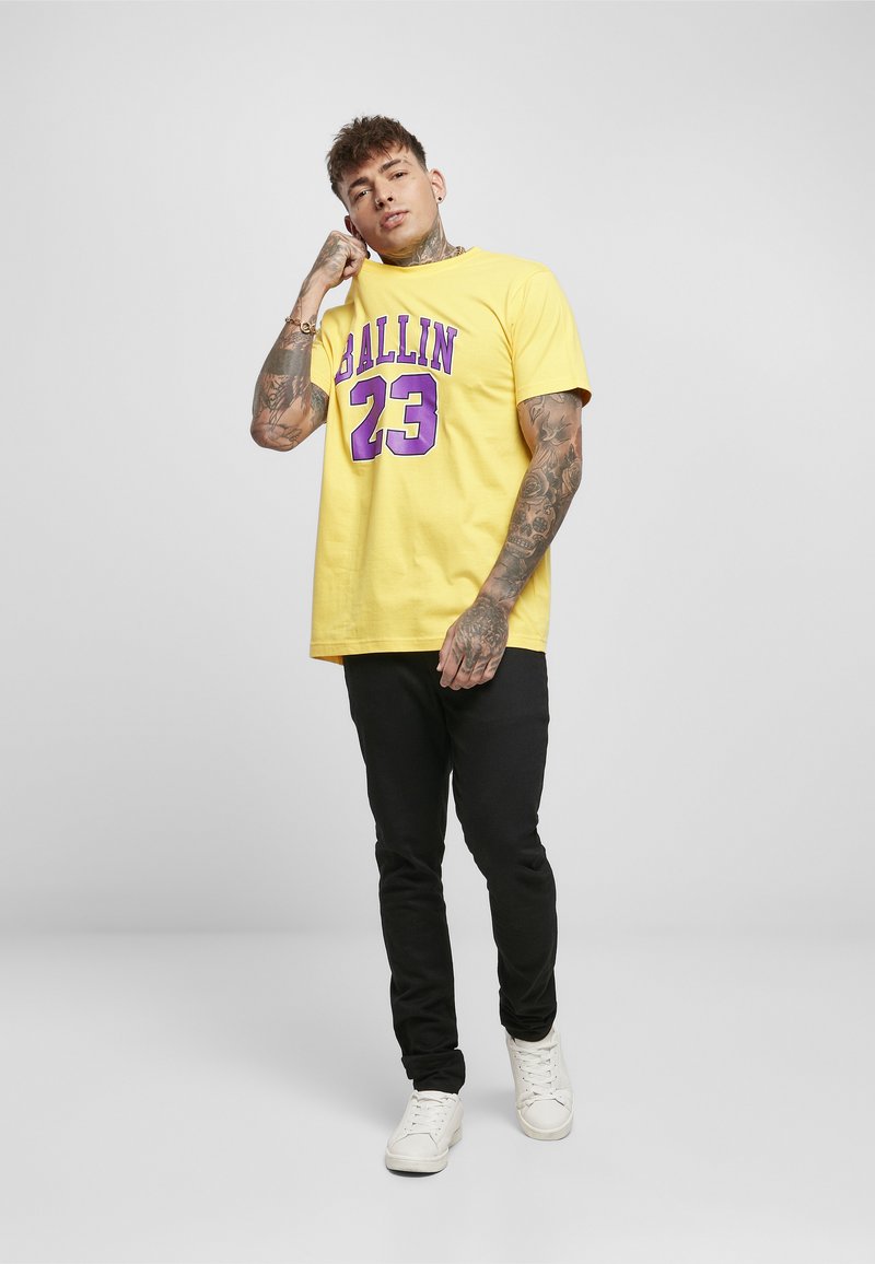 Mann mit Tattoos, der ein gelbes "Ballin 23" T-Shirt, schwarze Hose und weiße Sneaker trägt, steht und berührt seinen Nacken.