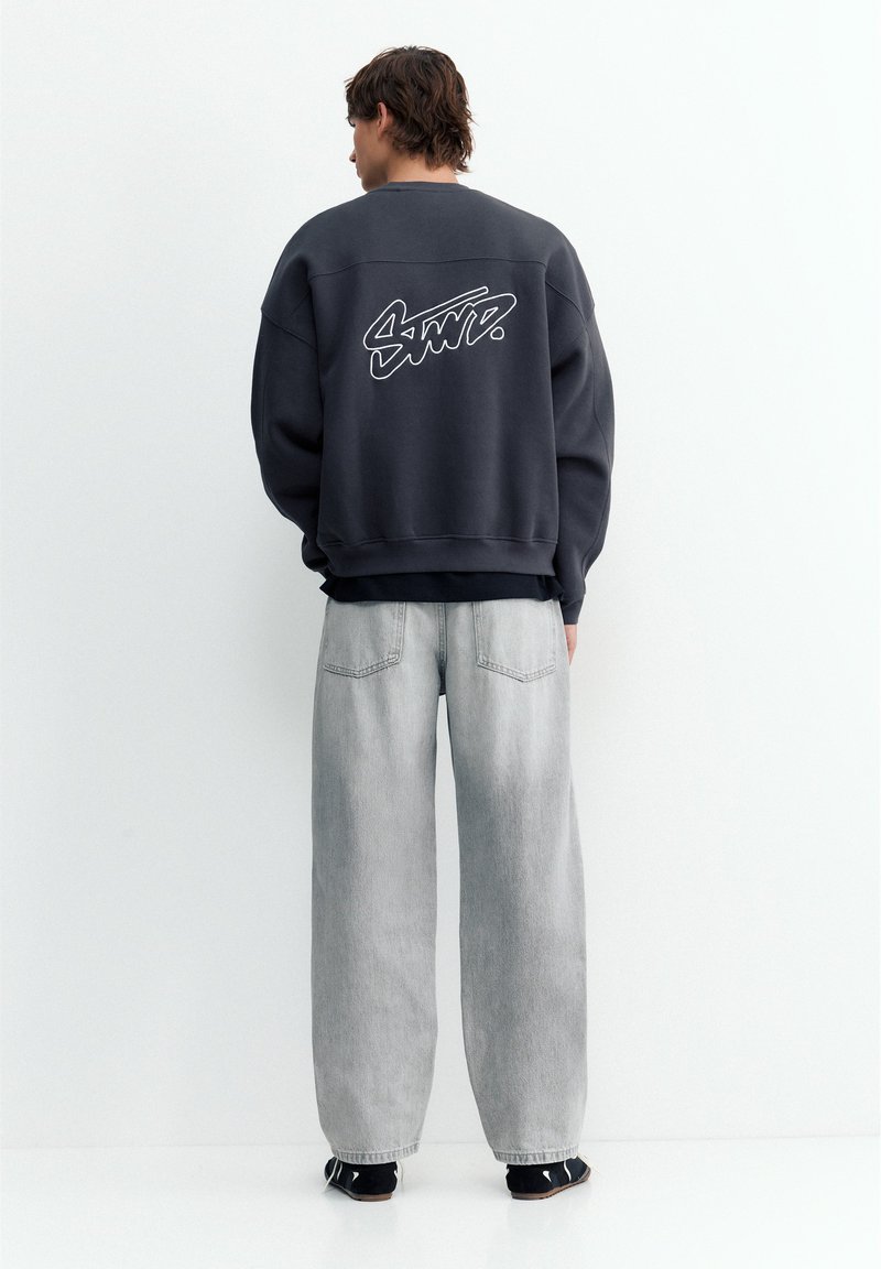 Jeune homme vu de dos, portant un sweat-shirt noir avec un motif blanc dans le dos et un jean ample gris clair sur fond blanc.
