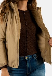 Giubbotto imbottito color sabbia con fodera in pelliccia artificiale, aperto per rivelare un pullover in maglia marrone con texture e motivi a rombi, decorato con bottoni marroni.
