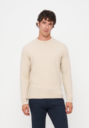 Homme aux cheveux bouclés foncés portant un pull beige clair à col rond et un pantalon bleu foncé, debout devant un fond blanc uni.