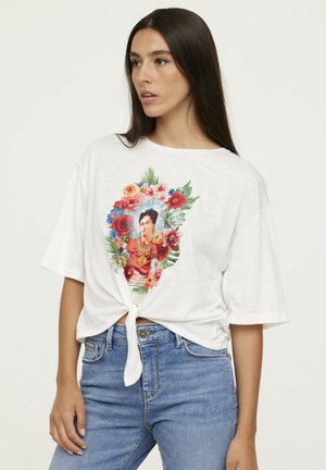 T-shirt blanc à manches courtes avec un grand motif floral coloré représentant un portrait au centre, noué à la taille, associé à un jean en denim.