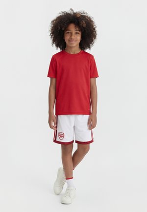 Kind mit lockigem Haar steht und trägt ein rotes Kurzarmshirt, weiße Sportshorts mit rotem Saum, weiße Socken und weiße Turnschuhe.