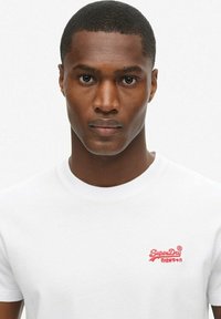 T-shirt bianca in cotone con un logo rosso sul lato sinistro del petto, collo rotondo e maniche corte, tessuto liscio.