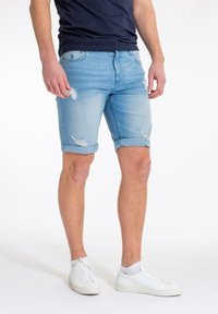 Mies, który ma na sobie jasnoniebieskie, przetarte szorty dżinsowe, białe niskie trampki i granatową koszulkę, stoi na tle białego tła.