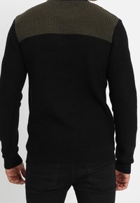 Schwarzer Strickpullover mit strukturierter Oberfläche, versehen mit einem einfarbigen dunkelgrünen Einsatz über den Schultern und langen Ärmeln.