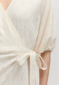 Wickelkleid aus strukturiertem hellbeigem Stoff, mit einem diamantartigen Spitzenmuster, kurzen Puffärmeln und einem gebundenen Taillenverschluss.
