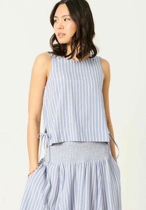 Woman wearing sleeveless blue and white vertical striped top with side ties and matching high-waisted skirt with smocked waistband.