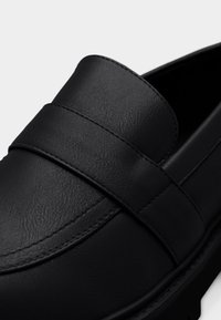 Μαύρο δερμάτινο μοκασίνι με λεία υφή, διακοσμητικό λουράκι στο πάνω μέρος και λεπτομερή ραφή κατά μήκος των ακρών σε λευκό φόντο.