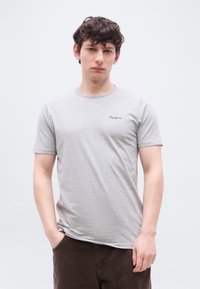 Νέος άνδρας με σγουρά σκούρα μαλλιά φοράει ανοιχτό γκρι μπλουζάκι Pepe Jeans και σκούρα παντελόνια, στέκεται με το ένα χέρι στην τσέπη μπροστά σε απλό φόντο.