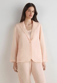 Světle růžový lněný blazer s klopami, zapínáním na jeden knoflík a dvěma předními kapsami, kombinovaný s odpovídajícím topem. Hladká textura.