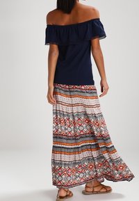 Kvinna som står, klädd i en marinblå off-shoulder topp och en lång mönstrad kjol med orange, svart och vita mönster, tillsammans med bruna sandaler.