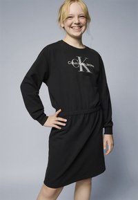 Vestido de sweatshirt preto com mangas longas, cintura elástica e logótipo Calvin Klein Jeans em prata. Textura suave e corte até ao joelho.