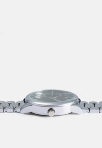 Sidabrinis metalinis laikrodis su apvaliu ciferblatu, juodu paviršiumi ir šviečiančiais valandų žymekliais. Yra tekstūruotas korpusas ir metalinė nuorodų apyrankė.