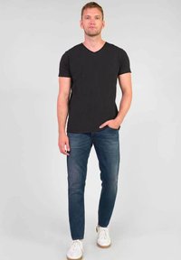 T-shirt nera a maniche corte con scollatura a V abbinata a jeans slim-fit blu e sneakers bianche; il modello posa su uno sfondo grigio.