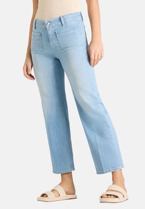 Vrouw draagt lichtblauwe rechte jeans, beige gebreide top en beige geweven slippers, staand tegen een effen achtergrond.
