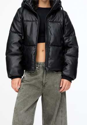 Personne portant une doudoune courte noire sur un crop top noir et un jean ample délavé gris-vert, debout devant un fond blanc.