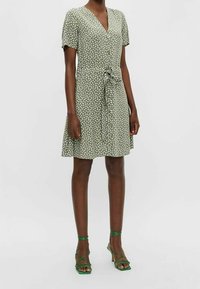 Robe verte à manches courtes avec des pois blancs, décolleté en V, taille marquée et détail à nouer. Associée avec des sandales vertes à lanières.