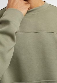 Sweatshirt vert olive en tissu texturé, doté d'un col rond et d'une couture horizontale traversant la poitrine pour un détail supplémentaire.