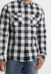 Camicia flanella a quadri bianca e nera con maniche lunghe, design con bottoni, due taschini sul petto e un'etichetta decorativa sul lato sinistro.