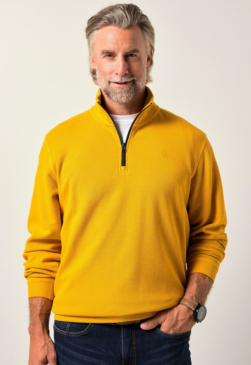 Uomo di mezza età con capelli e barba grigi, che indossa un maglione giallo a zip quarti e jeans blu, con una mano in tasca.