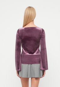 Lilla fløyelsgenser med lange ermer, bred utringning og klokkeformede ermer, kombinert med et grått plisset skjørt. Glatt, glinsende tekstur.