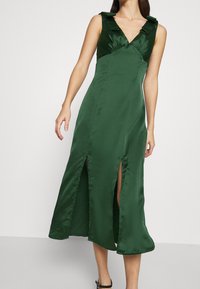 Vihreä satiininen midimekko, ilman hihoja, V-pääntiellä, solmukohtia olkapäillä ja edessä halkioita, päällä seisovalla henkilöllä.
