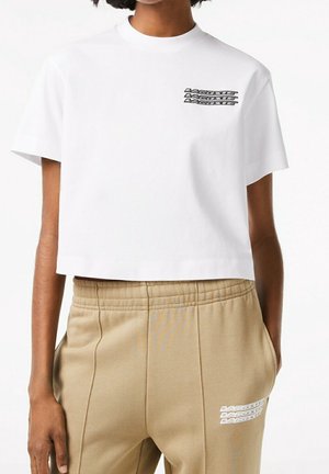 Osoba nosząca biały krótki t-shirt z czarnym napisem na piersi oraz beżowe spodnie z białym napisem przy kieszeni, ręce w kieszeniach.